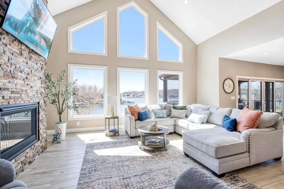 A large, comfortable sectional sofa sits facing a stone fireplace beneath expansive windows that fill the living room with natural light. The coffee table and tasteful decor invite gatherings, while the windows frame stunning views of the lake beyond.