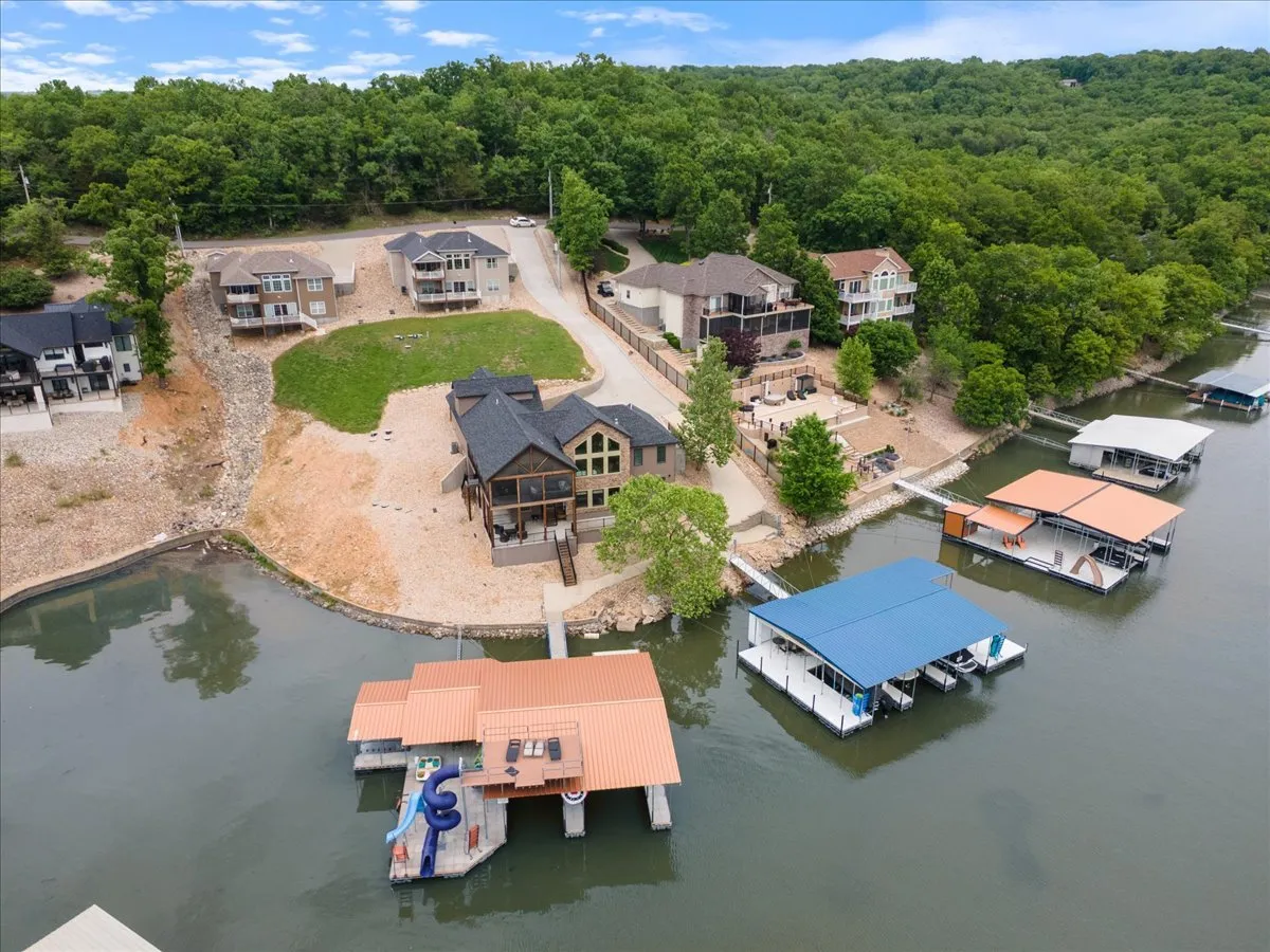 A broad view captures a modern waterfront home surrounded by lush greenery, with multiple docks and boats along the shoreline. The house appears welcoming and perfectly situated for lake adventures, while the boats and docks hint at seasonal recreational opportunities.