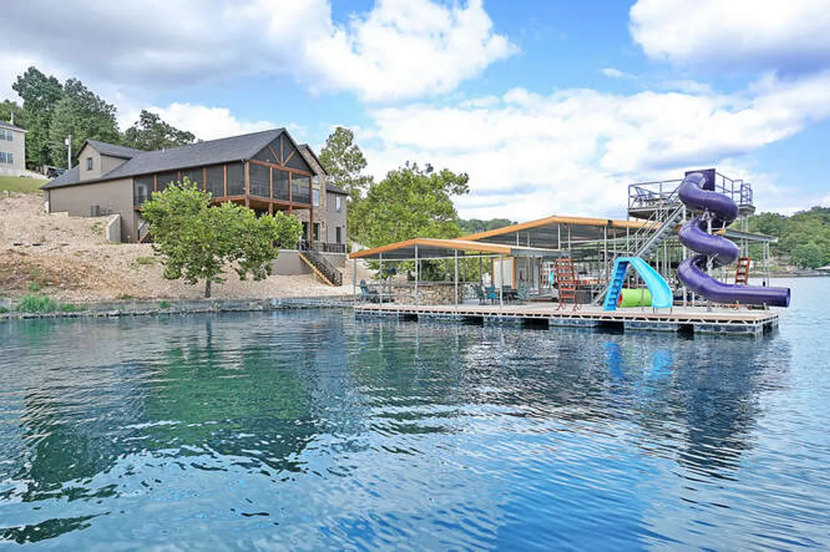 A wide shot of the property shows the house, multi-level dock, water slide, and the lake stretching out in the background. Children are seen sliding into the water, with boats docked nearby, and family members relaxing on the dock. The image conveys a blend of action, relaxation, and lakeside living.