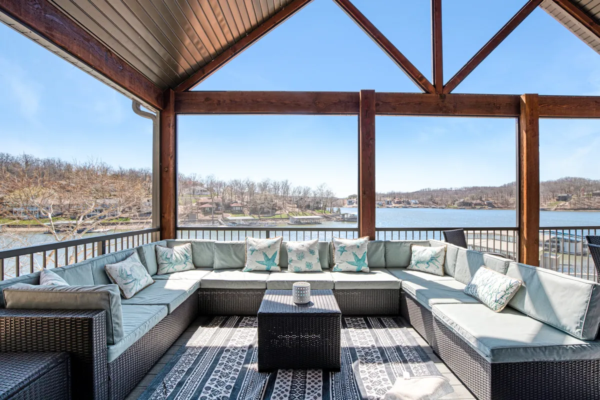 An outdoor sectional seating arrangement with decorative cushions sits atop a deck that overlooks a tranquil lake. A coffee table in the center suggests a perfect spot for morning coffee or evening cocktails. The scene is bathed in natural light and framed by beautiful lakeside views.