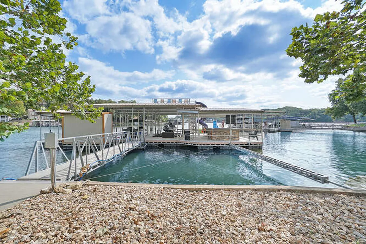 A sunny lakefront dock extends over calm water, surrounded by lush trees. Boats are moored alongside, and people can be imagined fishing, jumping into the water, or preparing for boating adventures. The inviting structure hints at spaces for swimming, sunbathing, and launching water sports.