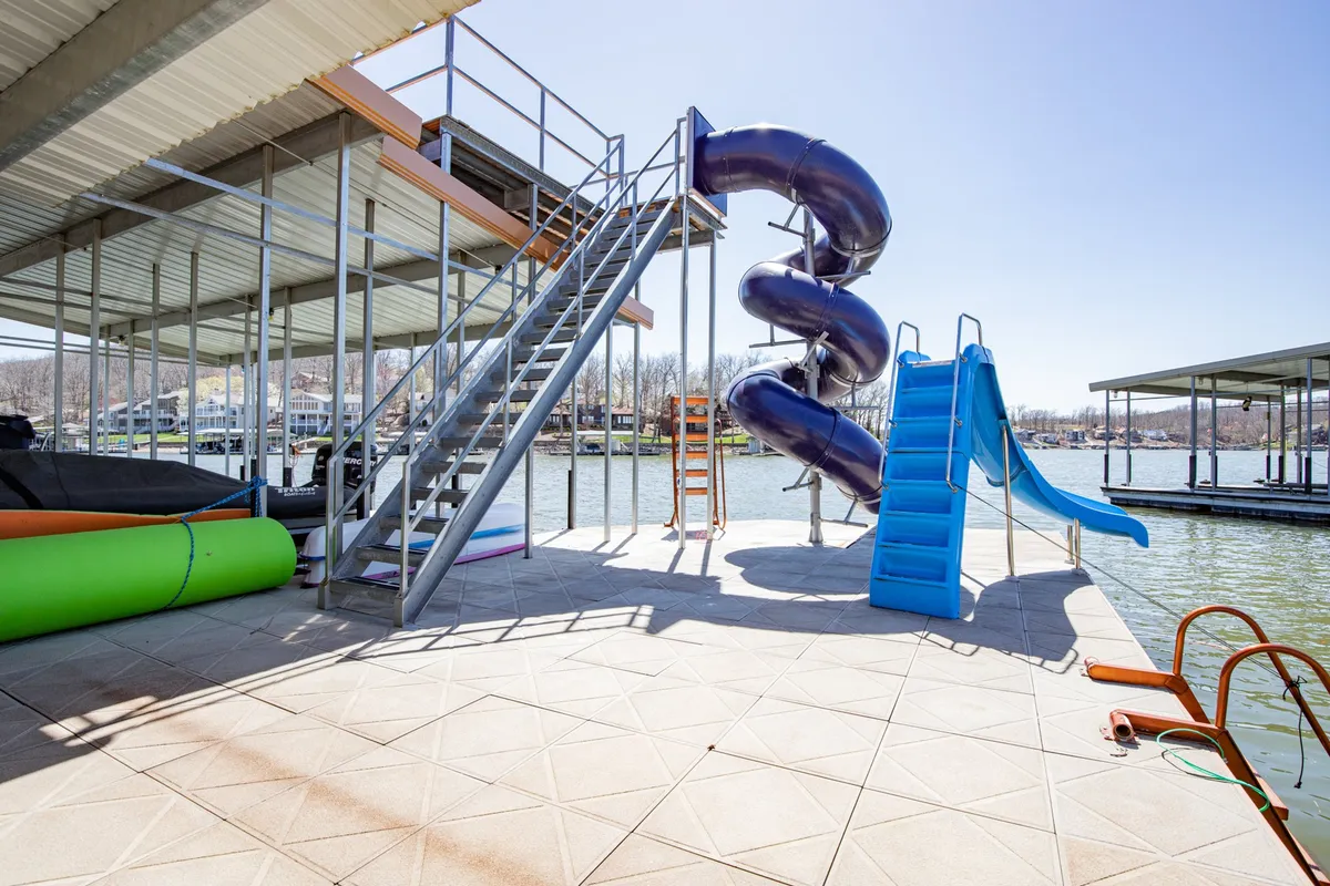 This exterior image focuses on a vibrant lakeside dock featuring a large, twisting water slide that plunges directly into the lake. Stairs connect different dock levels, while the sparkling swim area and playground equipment signal fun and activity. The scene is lively and sunlit, with people visible enjoying the water and the dock.