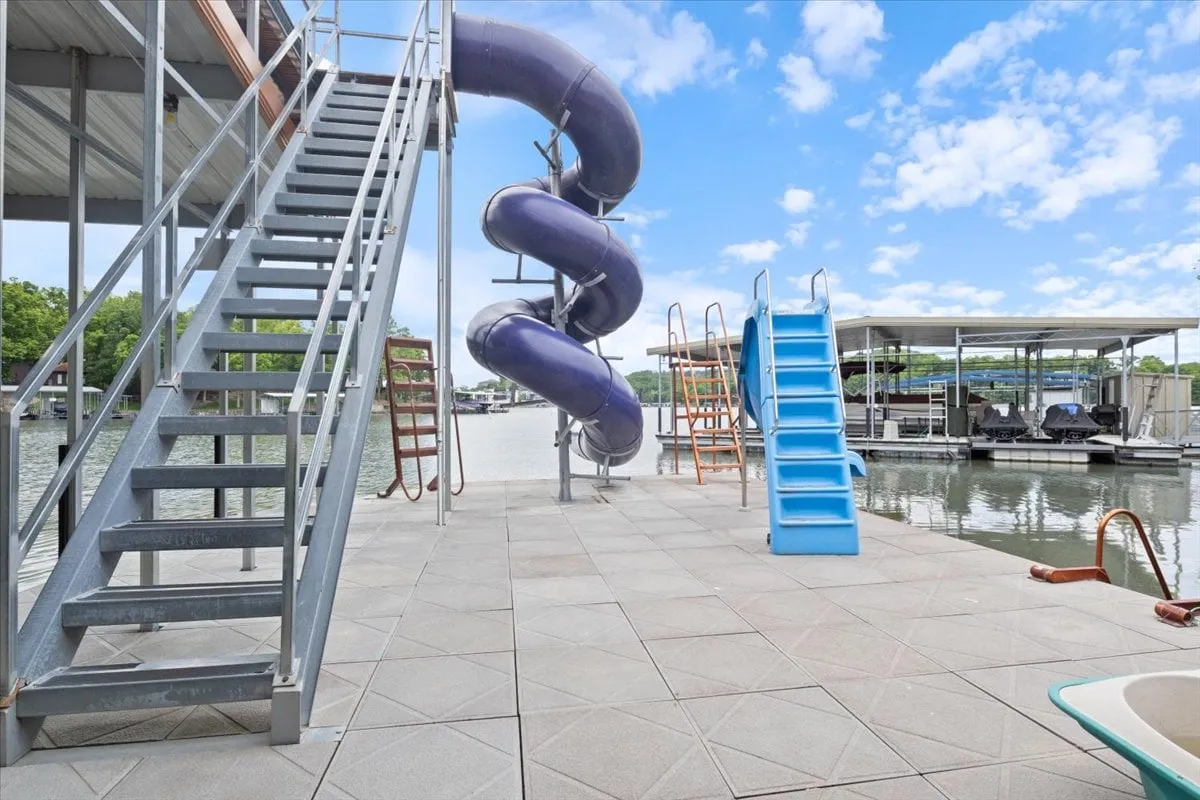 A playful dock scene highlights the water slide snaking down into the lake, a dedicated swimming area, and a staircase for easy access. The image captures people lounging near the water and others mid-slide, all enjoying the sun and water.