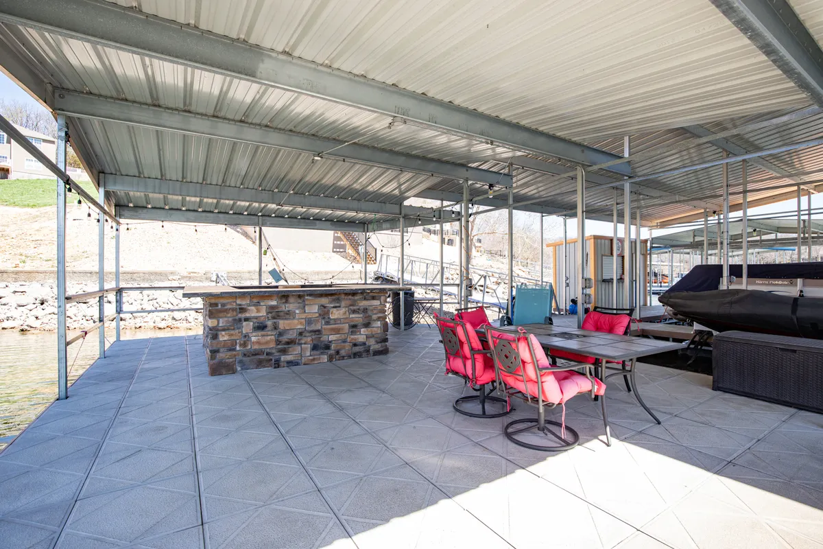 This image displays a stone bar area with bright pink chairs, a table for group activities, and a view of the boat dock and water beyond. The arrangement invites social gatherings, barbecues, and board games, with ample space for everyone to participate or spread out as desired.