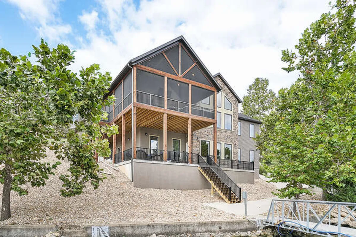 A spacious wooden porch wraps around a home with a beautiful stone facade and large, light-filled windows. The porch is surrounded by lush greenery, offering ample space for gatherings, relaxing, and enjoying expansive views of the lake. The scene radiates an inviting and serene mood, perfect for sunset views or morning coffee outdoors.