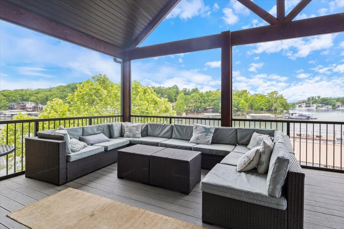 An inviting outdoor sectional sofa is positioned to enjoy panoramic views of the lake. Green trees frame the scene, and the tranquil water glistens in the background. The setting is perfect for relaxing outdoors in any season—whether sipping morning coffee or watching the sun set.