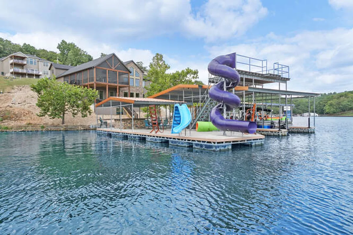 The image showcases the stunning lakeside exterior of a spacious luxury home in Linn Creek, Missouri, featuring a vibrant dock with an eye-catching enclosed tornado slide, a standard dock slide, a towering 20-foot jump platform, and inviting swim and sun areas. The lake sparkles in the background, framed by lush greenery, while the dock is clearly designed for family fun, water adventures, and relaxation. The scene radiates excitement, warmth, and the promise of unforgettable moments spent by the water.