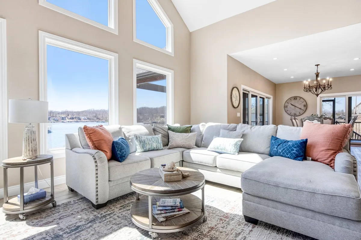 A spacious, light-filled living area features a large sectional sofa, a round coffee table, and wide windows that overlook tranquil lake waters. The space is styled for comfort and group gatherings, with decorative pillows and inviting seating.